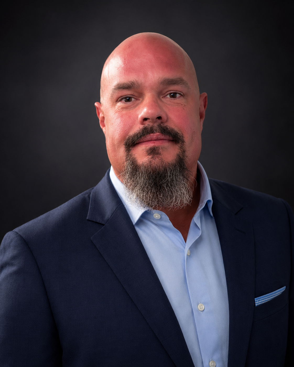 Studio headshot of a bald man with a grey-flecked beard, wearing a navy suit jacket over a light blue dress shirt, against a dark background.