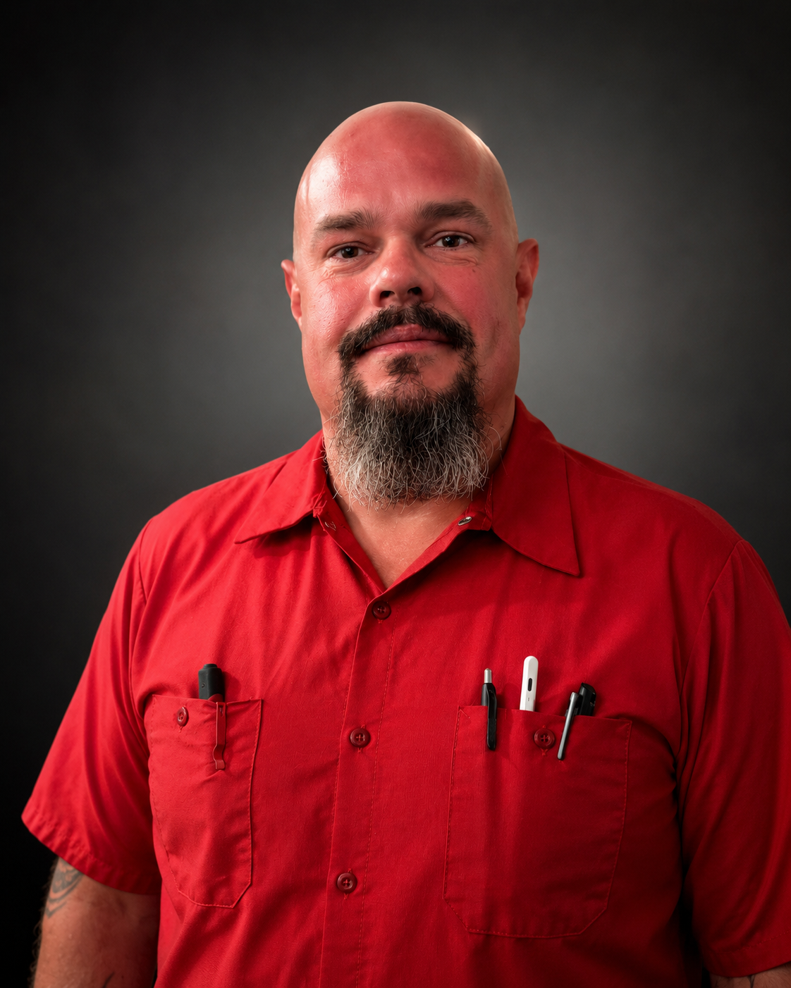 Studio torso shot of a bald man with a grey-flecked beard, wearing the red DHS Plumbing work shirt with pens clipped in the chest pocket, against a dark background. A tattoo is visible on the forearm.