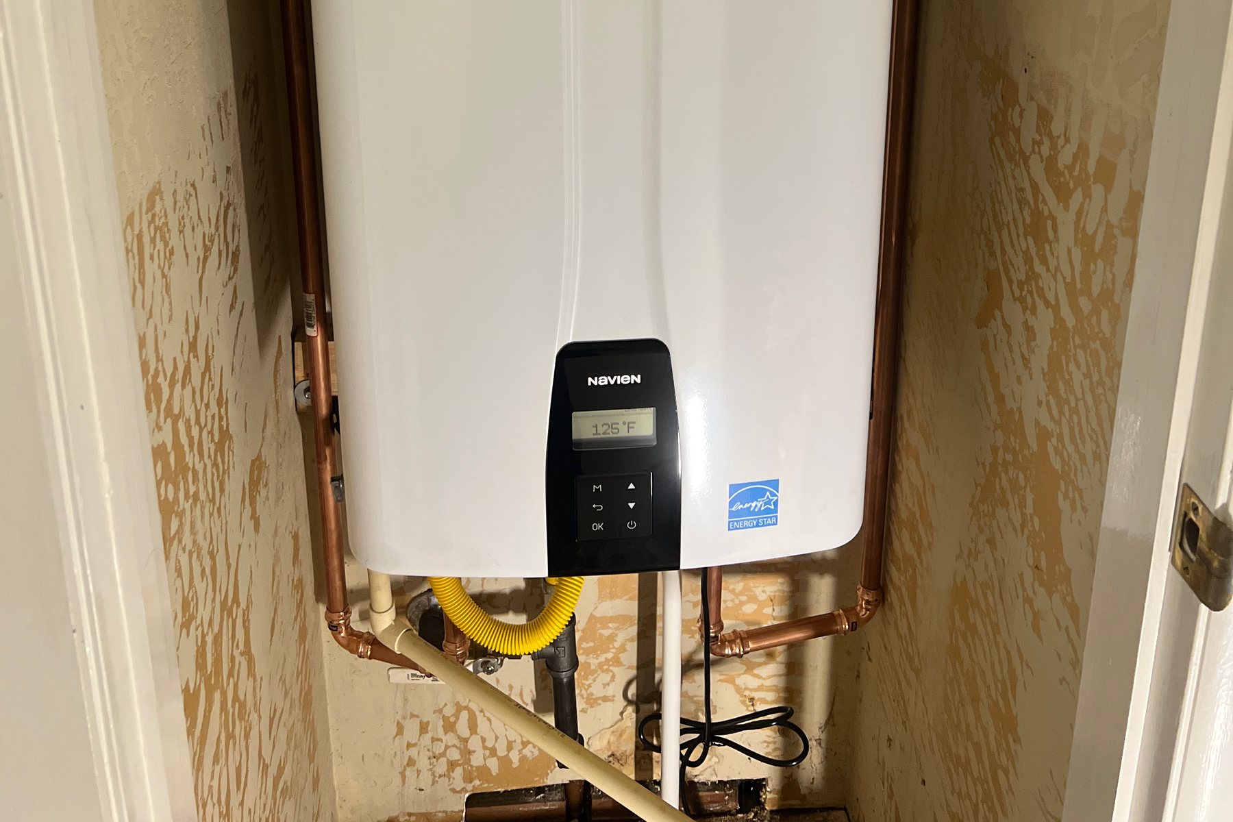 The gas connection under a wall-mounted Navien tankless: a yellow flex connector off black iron pipe, with copper supply lines running down either side