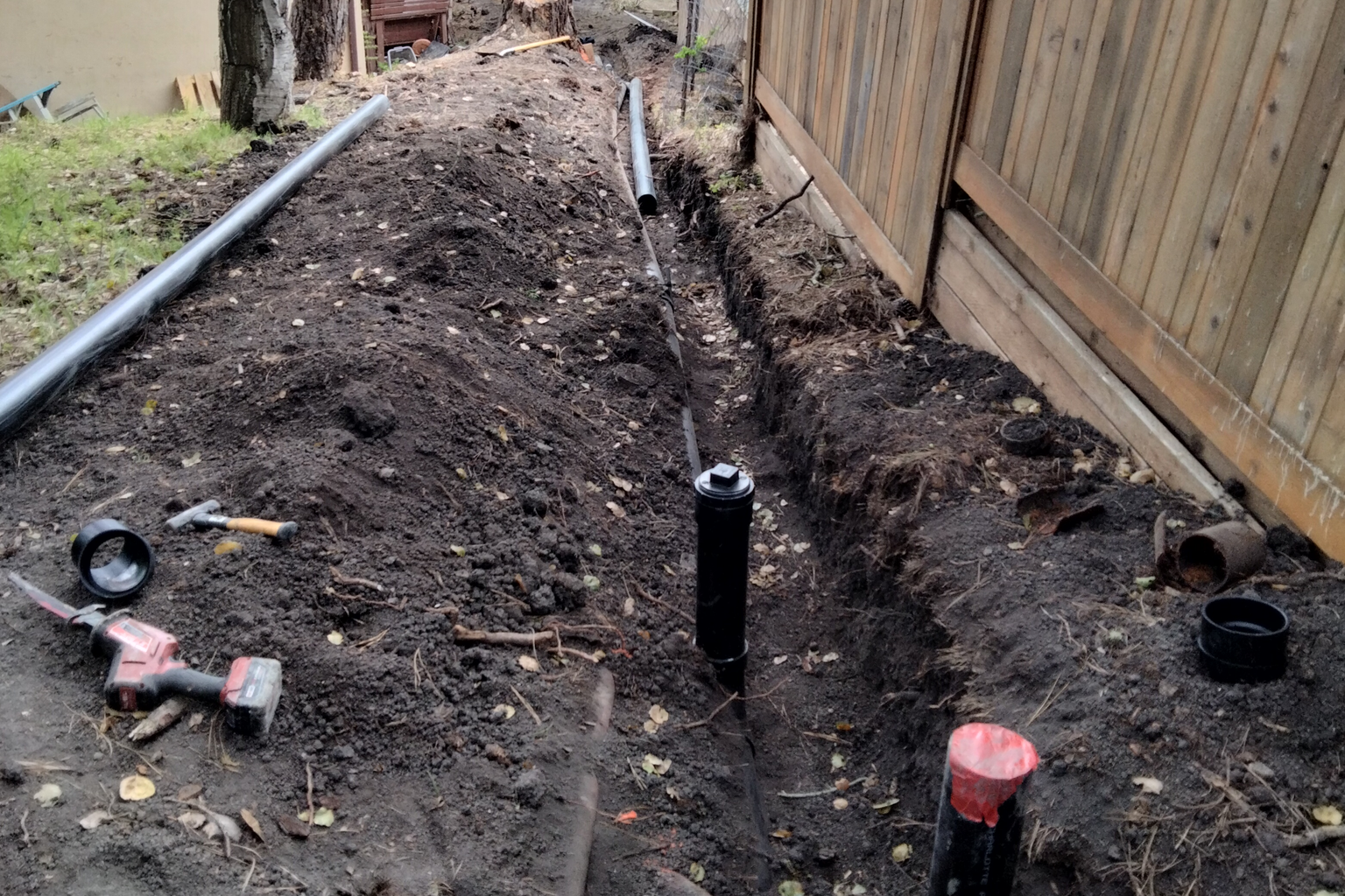 A replaced sewer lateral in a side yard, the trench backfilled along the fence line with new cleanout risers standing above grade