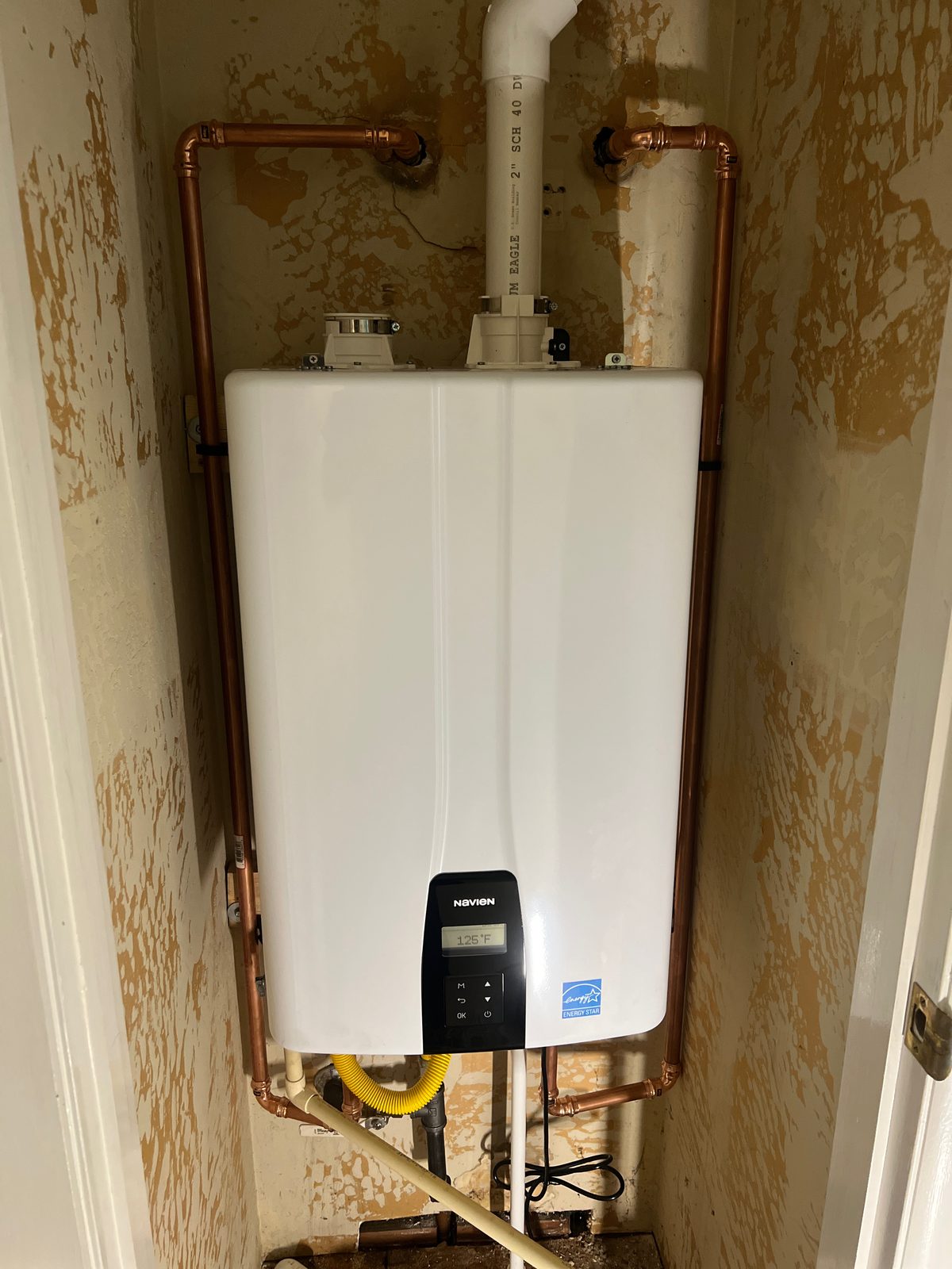 After: a new Navien tankless wall-mounted in the same closet, with new copper supply lines running up either side, a yellow gas flex connector below and a 2-inch schedule 40 vent out the top