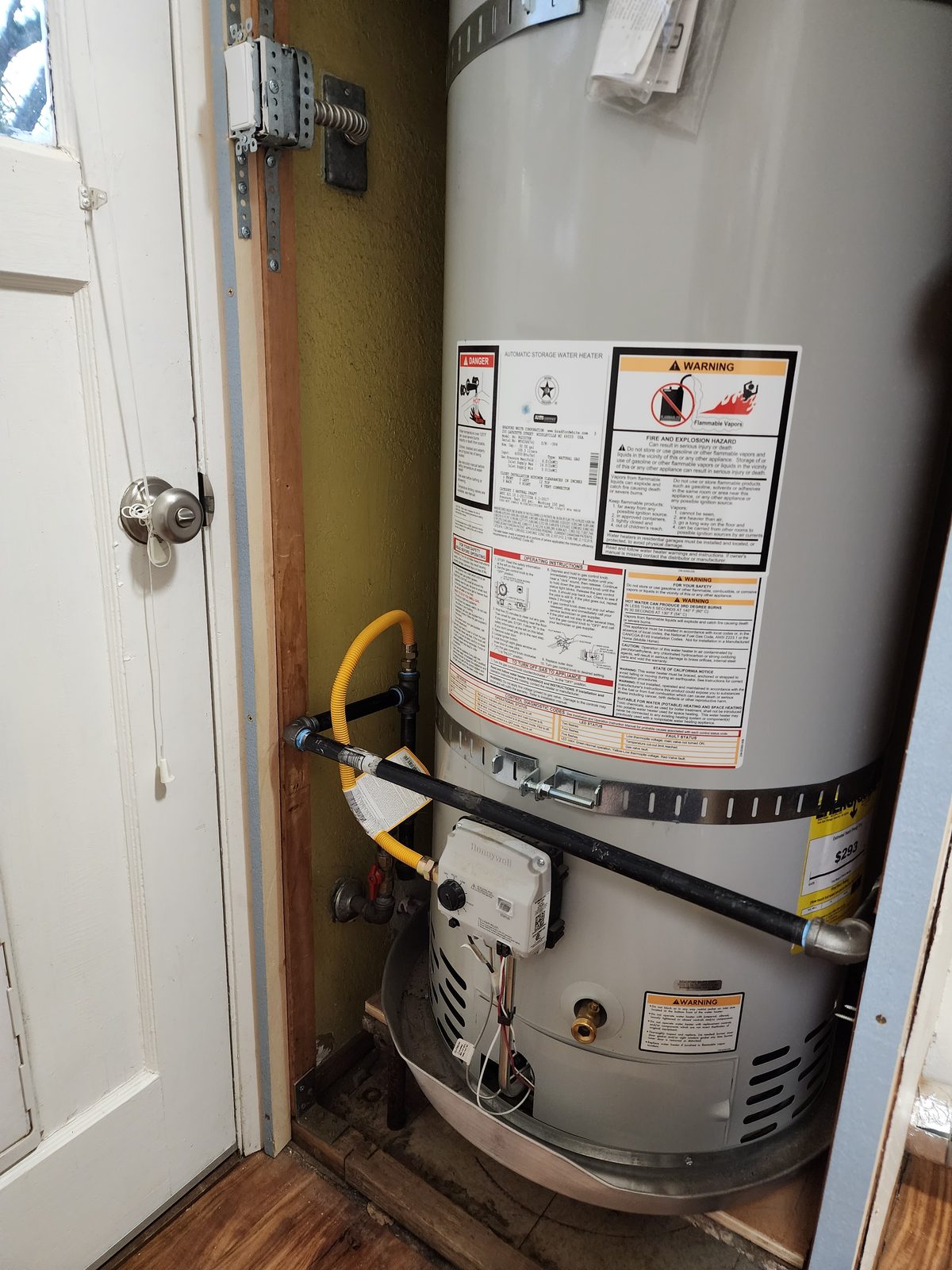 Before: the original gas tank water heater filling the closet, its warning labels facing out, with a black gas line, a yellow flex connector and seismic straps on the bare wall behind it